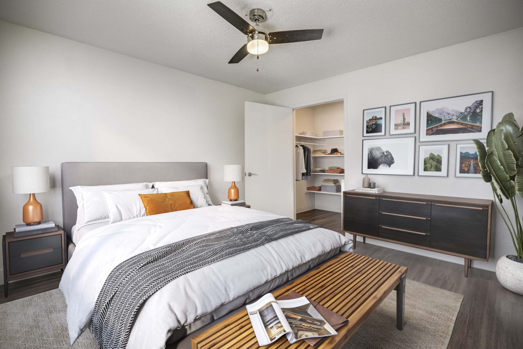 Bedroom with a dark bedside tables with lamps on them, an artificial plant in the corner, multiple wall arts and a closet with open door